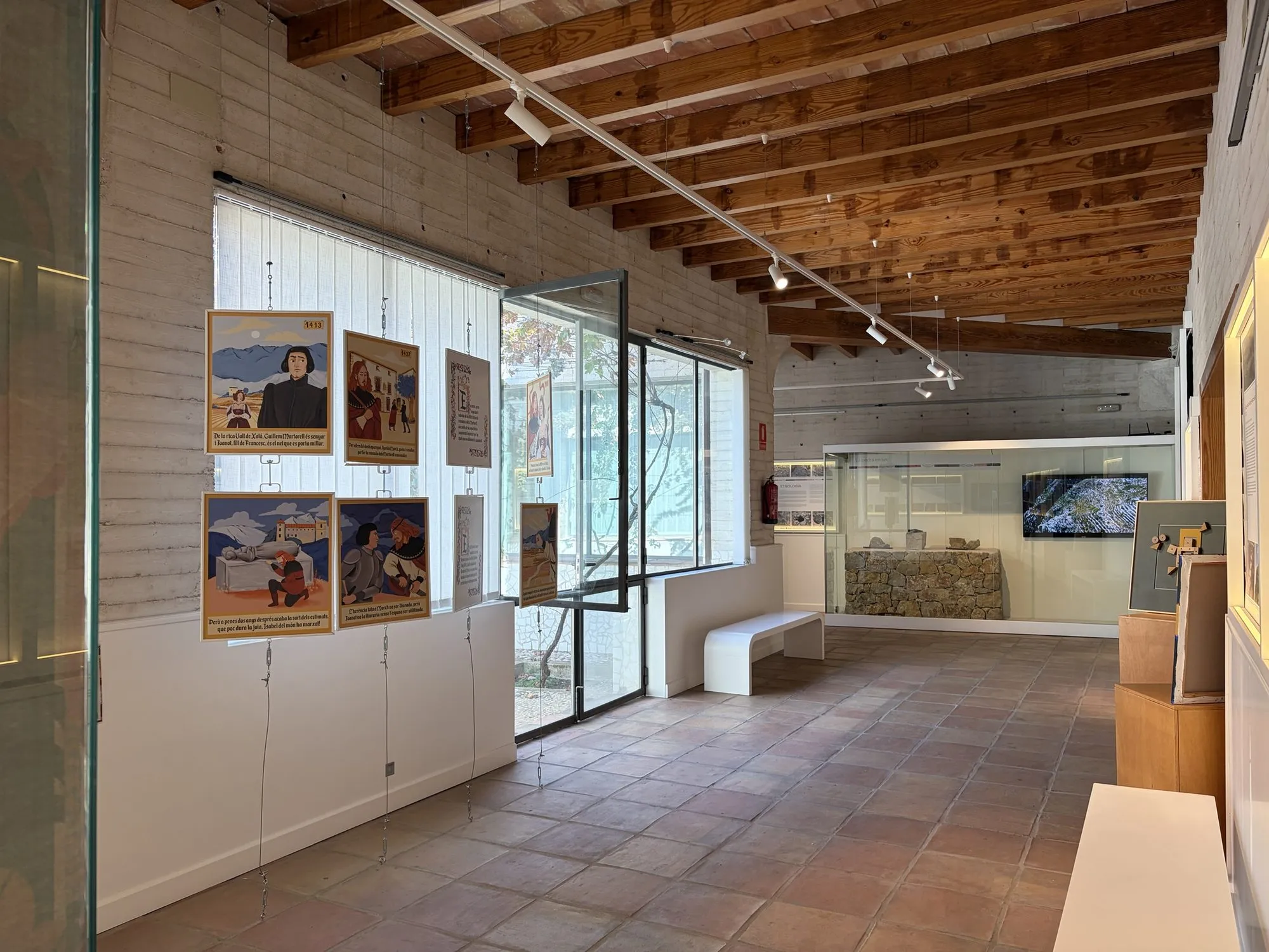 Room with a wooden beamed ceiling, illustrated panels on Joanot Martorell and, at the back, a display case with a dry-stone wall