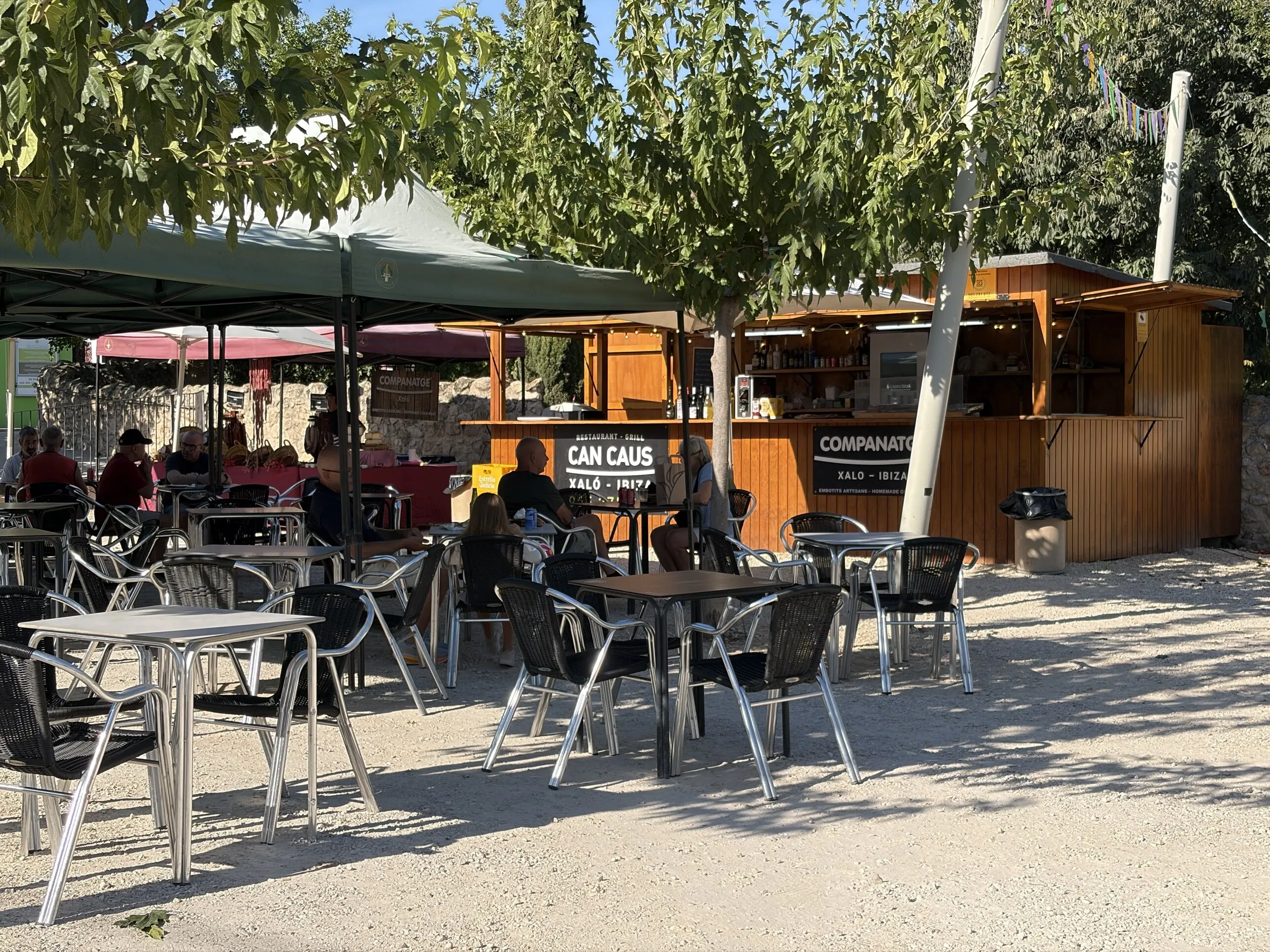 Tables and chairs in the shade by Companatge's fixed wooden stall, where people sit with a drink